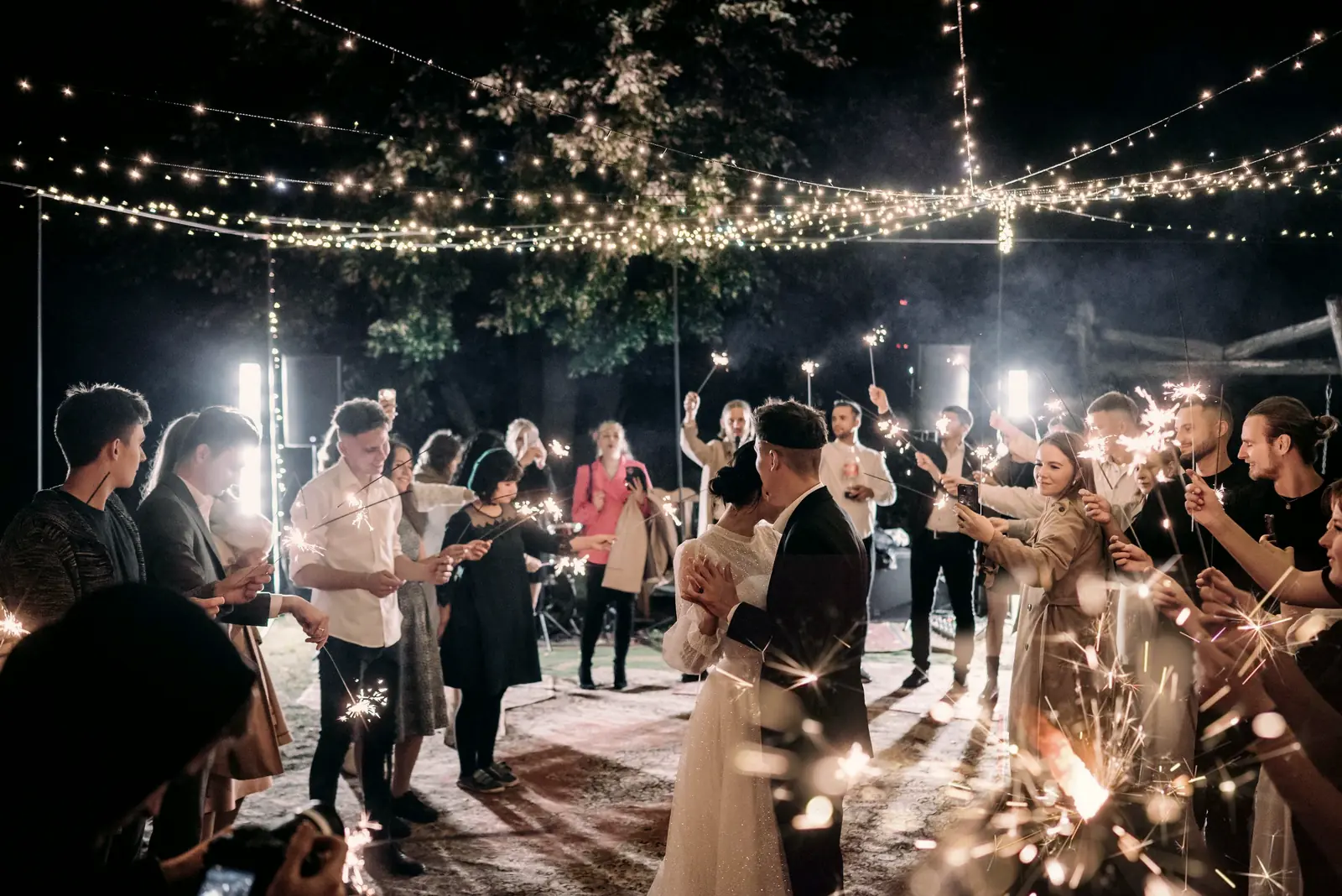 Ada and Theo slow-dance among guests holding sparklers under lights strung through the trees