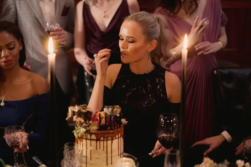 A guest tastes the wedding cake between tall black taper candles while friends in wine-red dresses watch