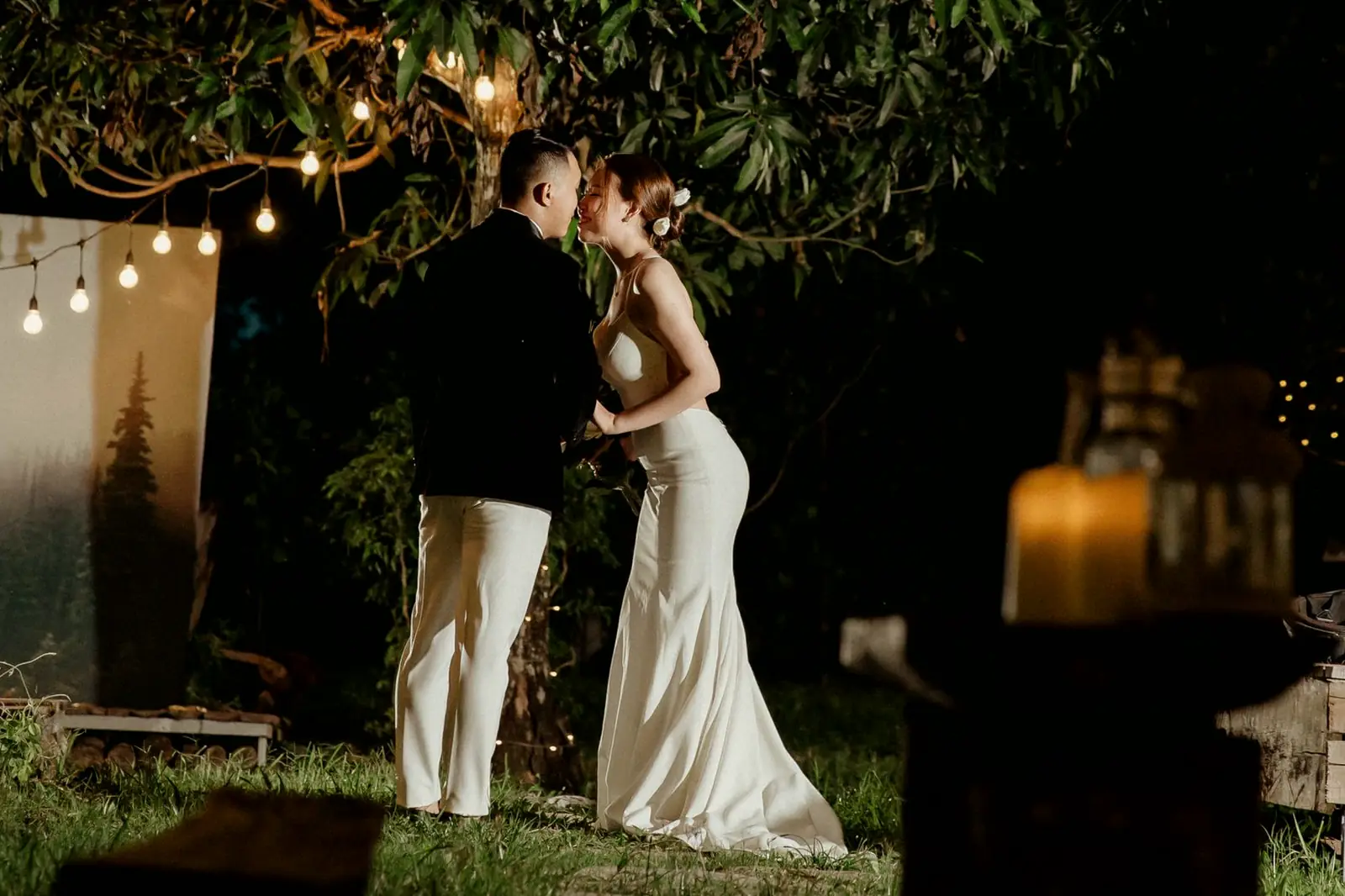 Lena and Sam kiss beside an old wooden barn under café lights and a big tree at night