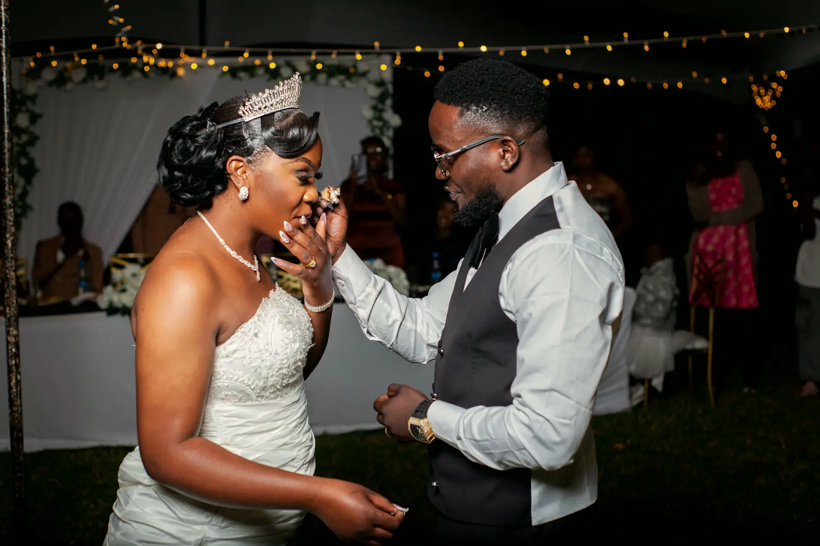 Nora laughs behind her hand as Eli feeds her cake under a tent strung with lights