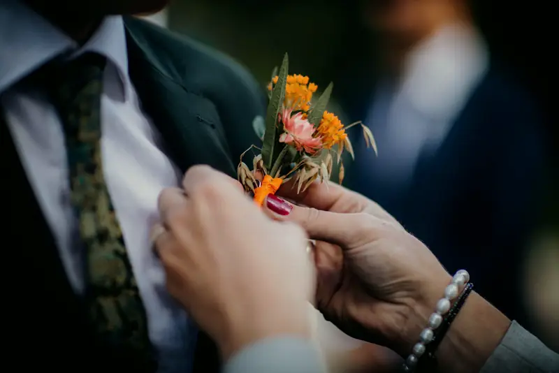 A woman’s hands pin a small orange boutonniere to a groom’s dark green lapel