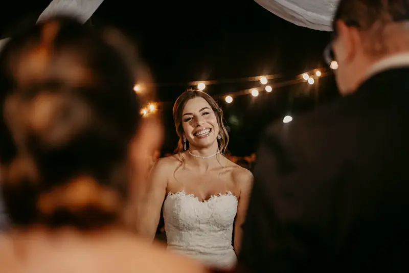 The bride smiles under strings of café lights as the best man speaks